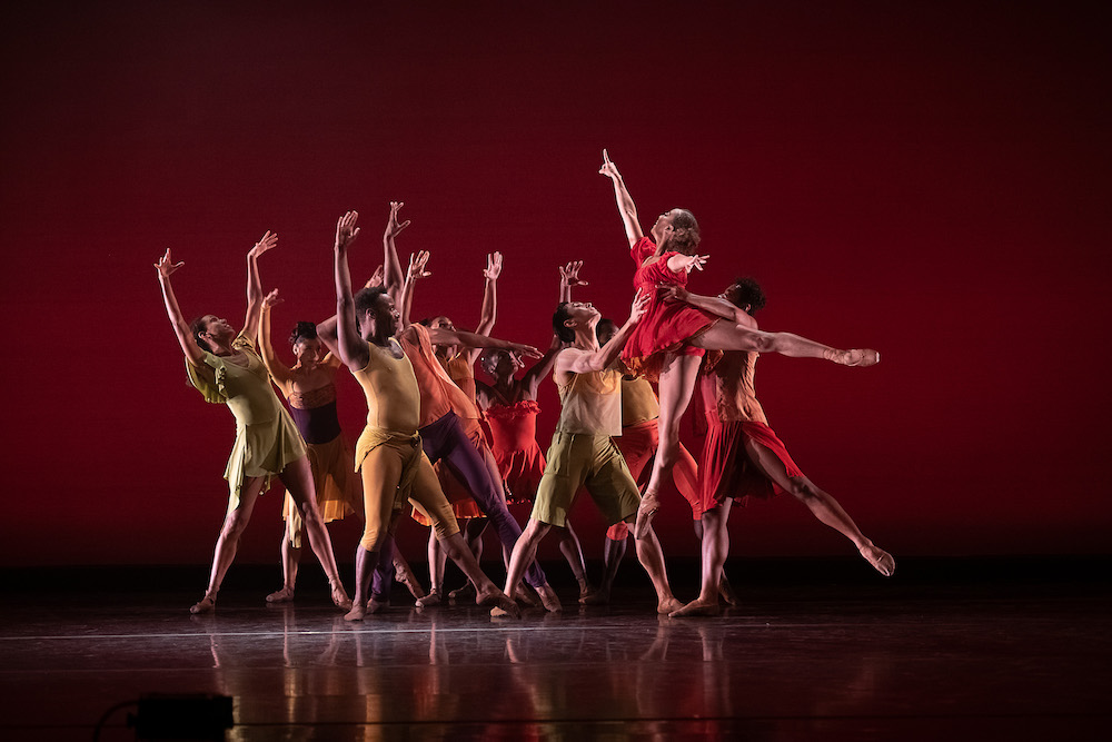 a woman in red leaps towards a company of dancers whose arms are raised in high spirited exultation. 
